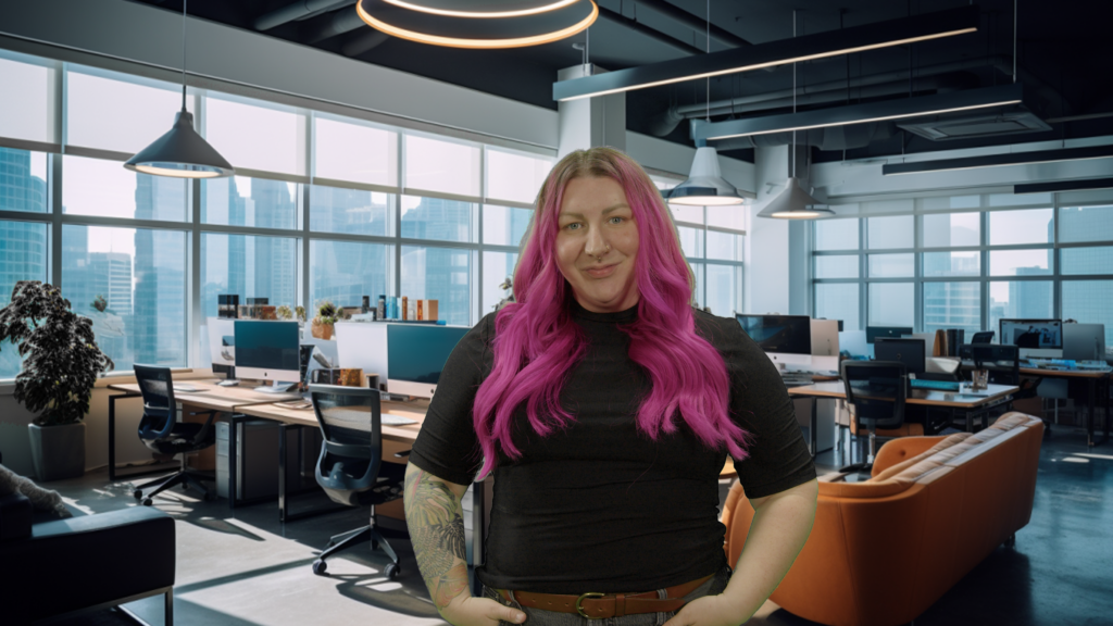 Sarah, with bright pink hair and a septum nose ring, stands confidently in the foreground of a contemporary office. They wear a black top and have visible tattoos on their arms. Behind them, the office is filled with modern furniture, computers on desks, and casual seating areas, all under stylish hanging lights. Large windows show a city skyline with skyscrapers, letting in natural light that brightens the room.