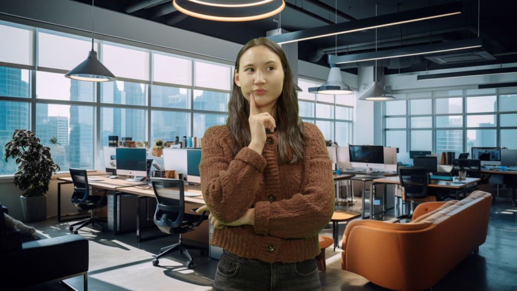 Alex stands in the foreground, her chin resting on her hand, as if deep in contemplation. She has long brown hair and is wearing a rust-colored cardigan over a black top, paired with dark jeans. The backdrop is a modern office space with large windows revealing a cityscape with blue skies. The office has a variety of workstations, some with computers, in a layout that promotes an airy and collaborative environment, illuminated by natural light and stylish overhead fixtures.