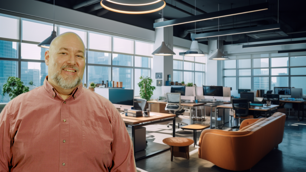 Will, with a bald head and a neatly trimmed beard, stands in the foreground of an open-plan office setting. He's wearing a salmon-colored button-down shirt. The office space behind him is well-lit with natural light streaming through large windows that offer a view of urban high-rises. The area is outfitted with modern desks, computers, comfortable office chairs, and communal couches, all under chic pendant lighting. The atmosphere conveys a professional yet relaxed work environment."