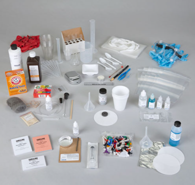 Various laboratory equipment and supplies laid out on a light gray surface. The items include gloves, test tubes in a rack, a test tube brush, pipettes, a bottle of hydrogen peroxide, boxes labeled 'Universal Indicator' and 'Magnesium Ribbon', petri dishes, a beaker, plastic bottles with nozzles, cotton swabs, a small bottle labeled 'Iodine Solution', a wire brush, several markers, and a roll of aluminum foil. These supplies are typically used in educational science experiments or classroom demonstrations.