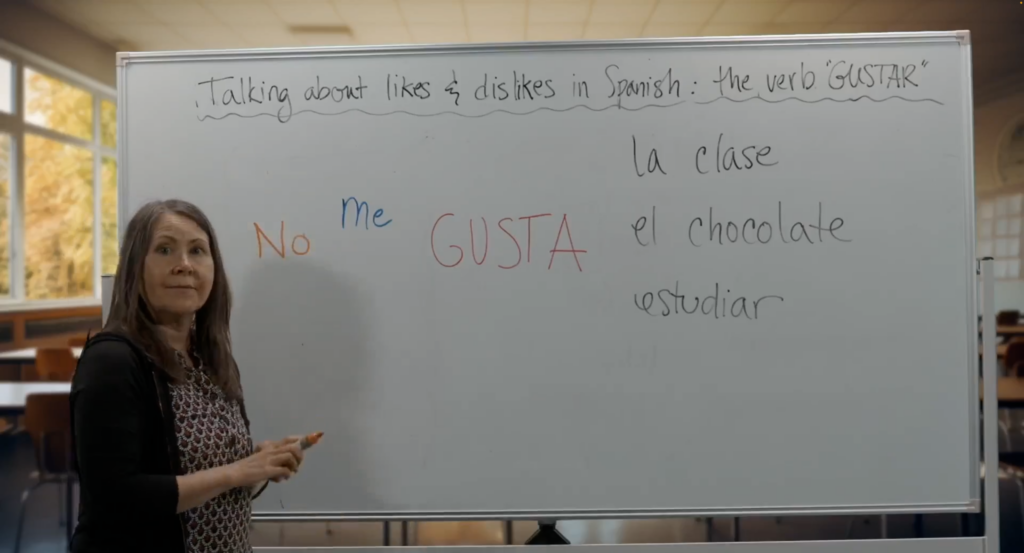 The image shows a woman standing in front of a whiteboard in a classroom, giving a lesson on expressing likes and dislikes in Spanish using the verb "gustar." The top of the whiteboard has the heading: "Talking about likes & dislikes in Spanish: the verb 'GUSTAR'" written in English. Below that, the phrase "No me gusta" is written in colored markers, with "No" in orange, "me" in blue, and "GUSTA" in red. To the right of the phrase, there are examples of objects and activities one might like or dislike, written in black: "la clase" (the class), "el chocolate" (chocolate), and "estudiar" (to study). The woman appears to be engaged in teaching, holding a marker and looking toward the camera. Behind her, large windows reveal an outdoor scene with trees showing autumn foliage.