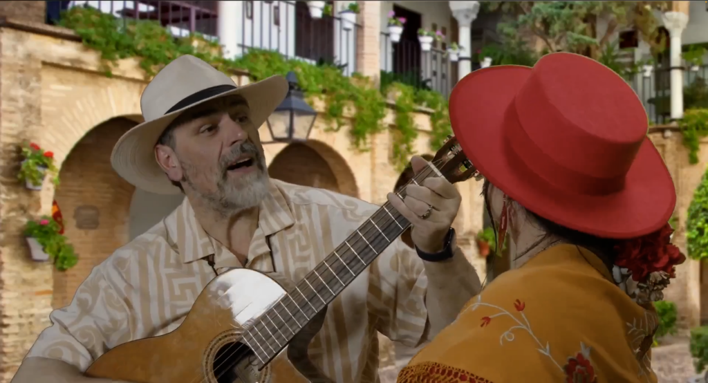 The image depicts a man, José García-Paine, and a woman, his wife Sofía, in traditional attire, performing music together. The man, with a beard and wearing a light-patterned shirt and a beige fedora, is strumming a guitar and singing. The woman, partially visible from behind, is wearing a bright red wide-brimmed hat and a yellow shawl with red floral patterns. They appear to be in an outdoor setting, with a backdrop of a rustic building adorned with green plants and arched doorways, giving the scene a festive and cultural ambiance.