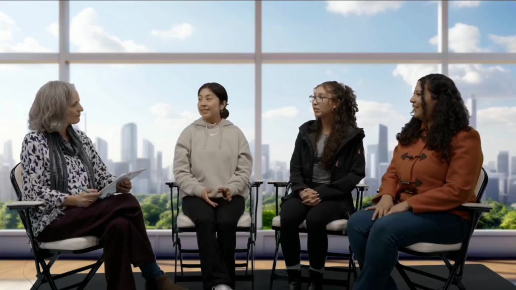 The image shows a group of four women seated in a discussion setting. Three younger women, dressed in casual outfits (hoodies, jackets, and sweaters), are sitting side by side, facing an older woman who is holding a clipboard. The older woman is dressed in a patterned blouse with a scarf, actively engaged in the conversation. Behind them, large windows showcase a city skyline with tall buildings and clear blue skies, indicating a modern, indoor space with a bright and open atmosphere.