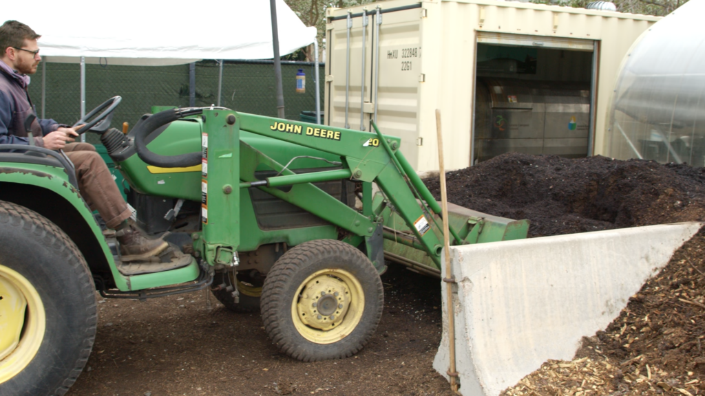 A person operates a John Deere tractor to move a large pile of compost at an outdoor composting facility.